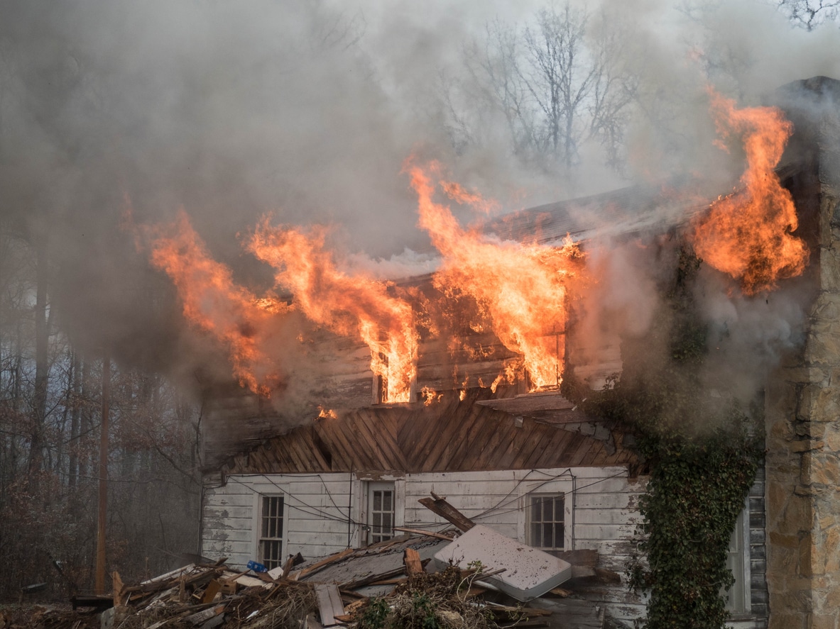 A severely damaged house engulfed in flames, with large orange fires and thick gray smoke pouring from the roof and upper floors.